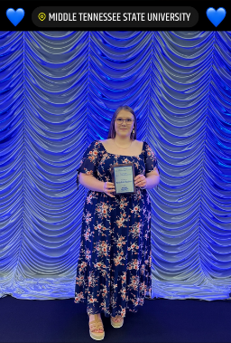 A female student in a flowered dress holding an award plaque, standing in front of a fancy blue curtain background. Header at the top says Middle Tennessee State University in white on a black background, with blue hearts.