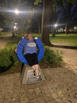 A female student sitting on the middle of the Blue Horseshoe with arms wrapped around the outside. Wearing a blue MTSU t-shirt and black capri leggings. The background is Walnut Grove at night.