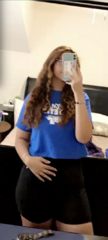 A female student taking a selfie in a mirror, with the phone in front of her face. Wearing a blue MTSU t-shirt and black shorts. Background is a bedroom.