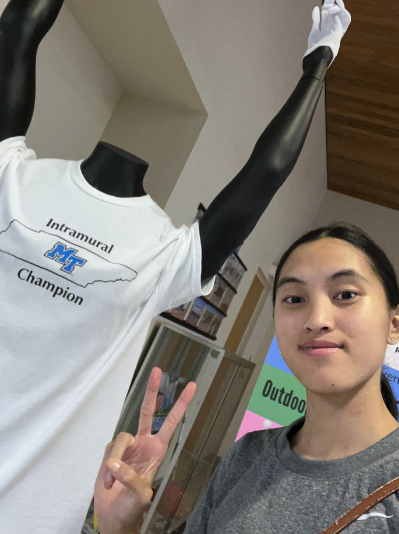 A female student holding up two fingers in a peace sign, wearing a gray t-shirt. Standing by a mannequin with arms raised in a victory sign, wearing a white Intramural Champion t-shirt.