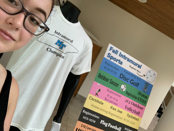 A female student wearing glasses and a black tank top. Standing in front of the mannequin, which has his arms by his sides this time. Fall Intramural Sports sign in the background, with details about intramural events this semester.