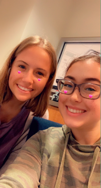 Two female students smiling for a selfie. One is wearing a camo hoodie and glasses. The other is wearing a purple shirt. They both have virtual heart emojis on their cheeks.